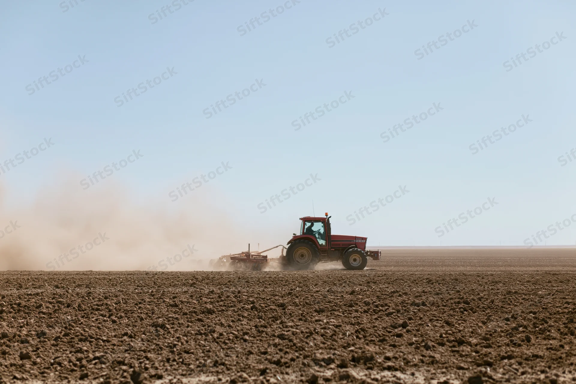Red tractor pulling a tiller across a dry farm field kicking up dust under a clear sky