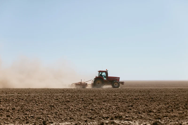 Red tractor pulling a tiller across a dry farm field kicking up dust under a clear sky