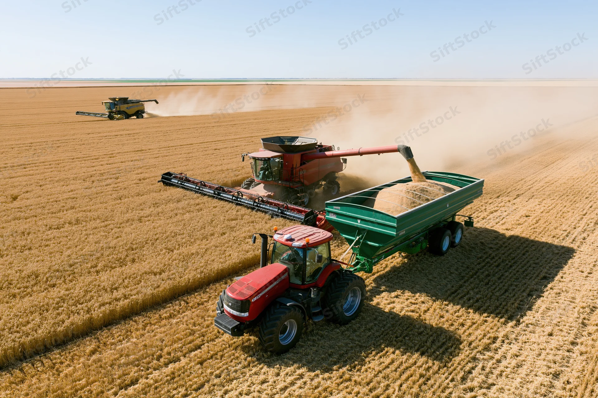 Red tractor with green grain trailer parked in foreground while combine harvester unloads wheat into trailer in golden field