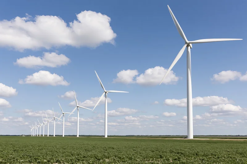 Row of white wind turbines receding in perspective across a green field under a blue sky with scattered clouds