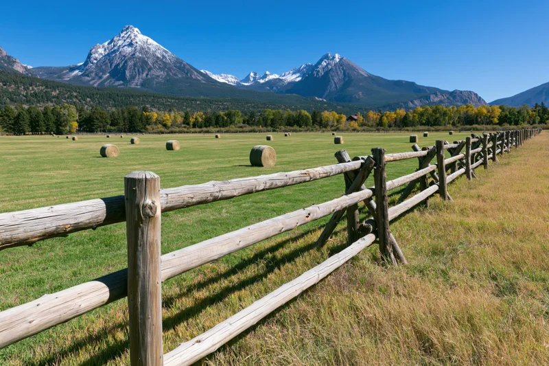 Wooden fence running diagonally across a green pasture with hay bales and snow-capped mountains in the background