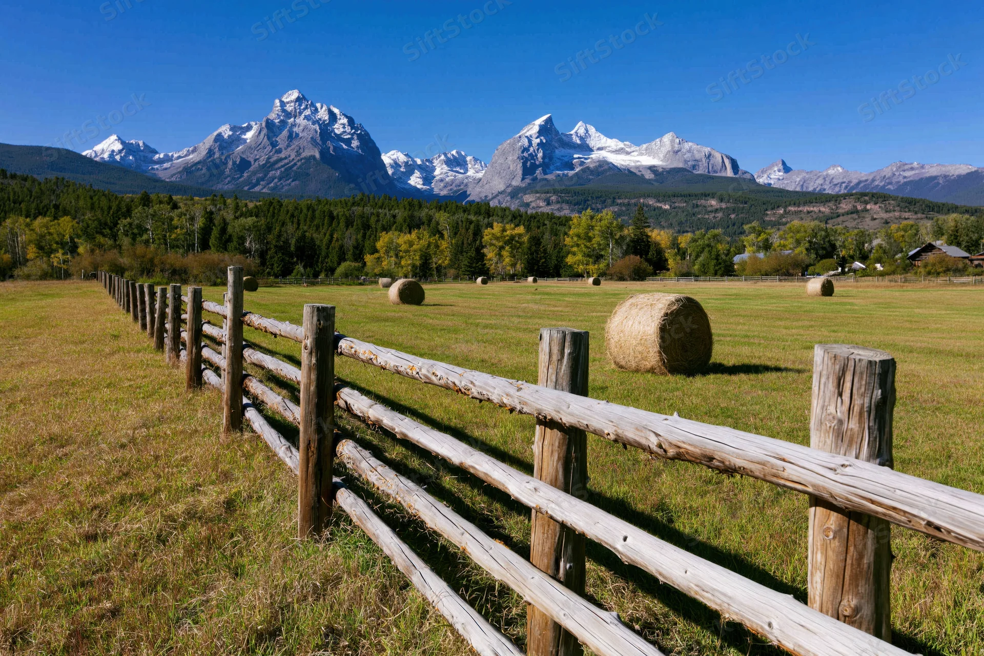Wooden fence running diagonally across a grassy field with hay bales and snow-capped mountains in the background