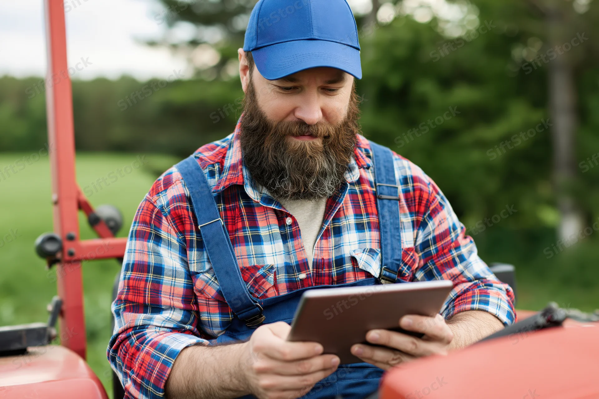 Bearded man in overalls using tablet on tractor in field