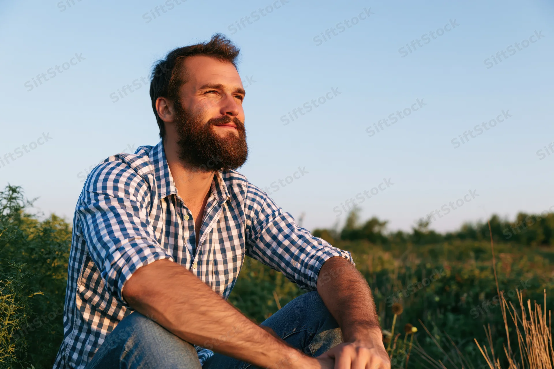 Bearded man sitting in a field during golden hour, wearing a checkered shirt and jeans, looking off into the distance with a relaxed smile