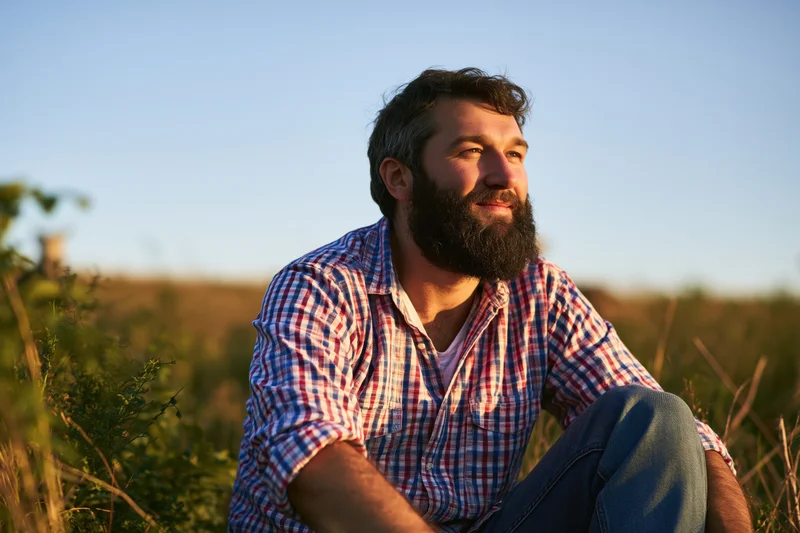 Bearded man sitting in a field at sunset wearing a checkered shirt and jeans