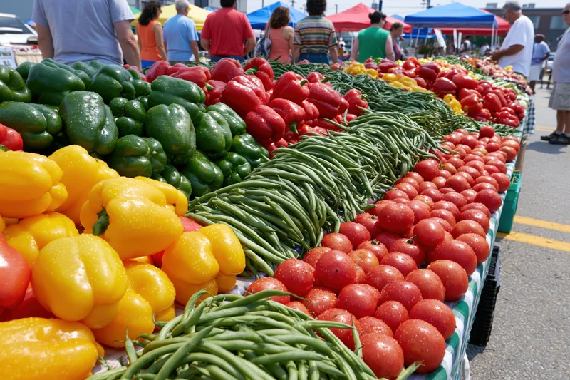 Rows of fresh vegetables including peppers, tomatoes, and green beans displayed on a table at an outdoor farmers market
