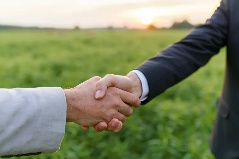Two men shaking hands in a green field at sunset