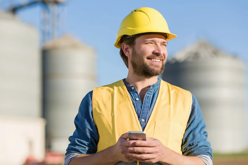 Construction worker in yellow safety helmet and vest holding smartphone outdoors at industrial site