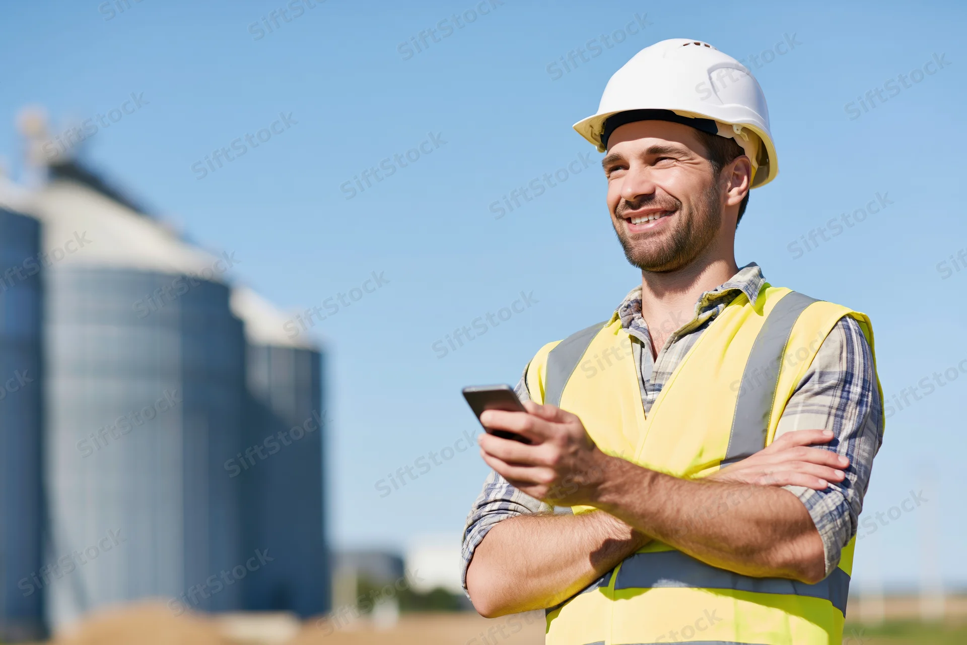 Construction worker smiling while holding smartphone with arms crossed outdoors at industrial site