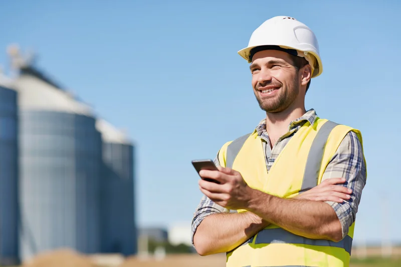 Construction worker smiling while holding smartphone with arms crossed outdoors at industrial site