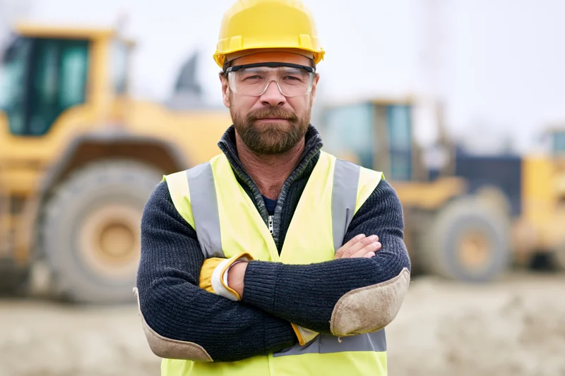 Construction worker with arms crossed wearing safety gear on a construction site