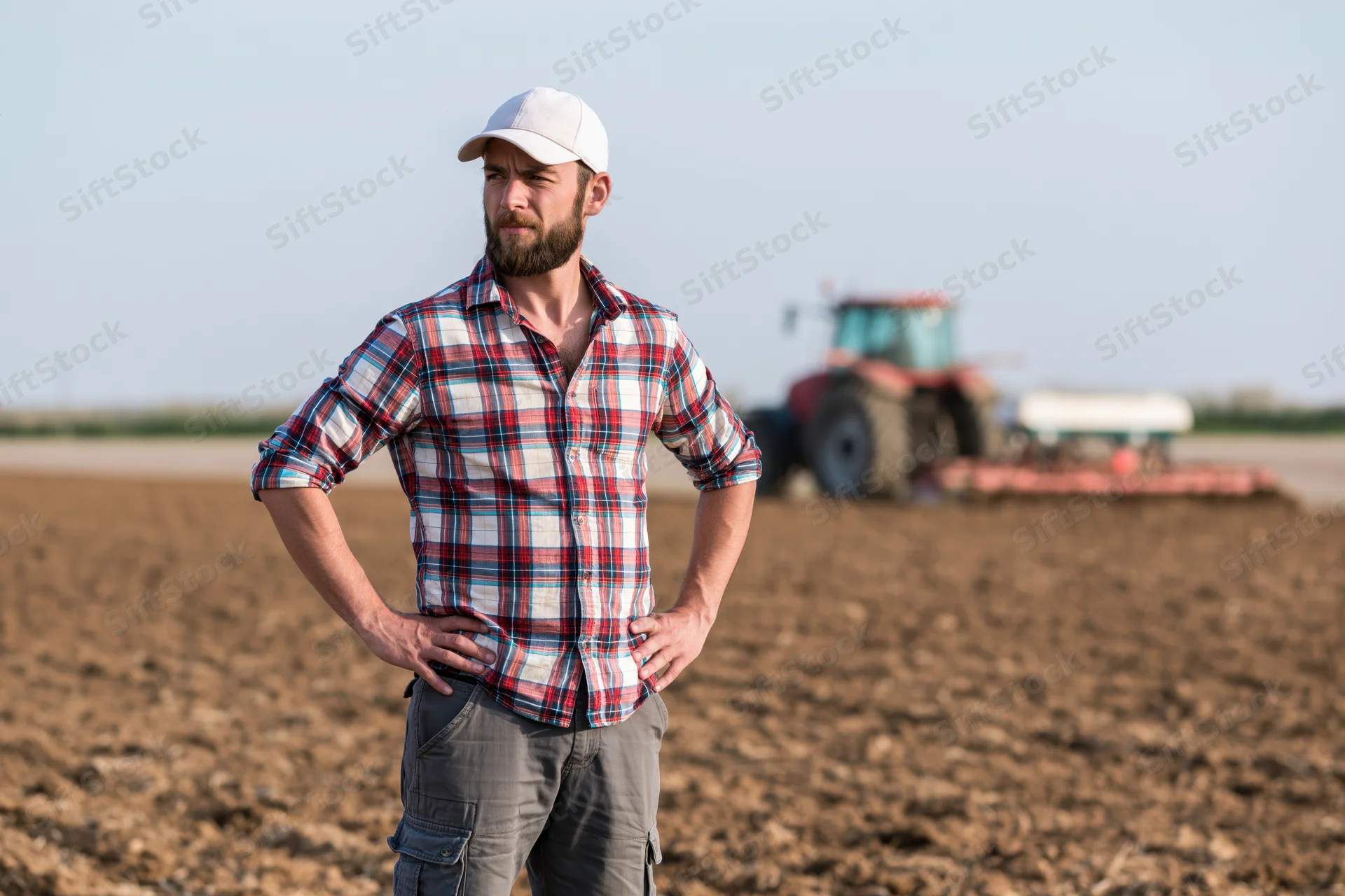 Farmer standing with hands on hips in a plowed field with tractor in background