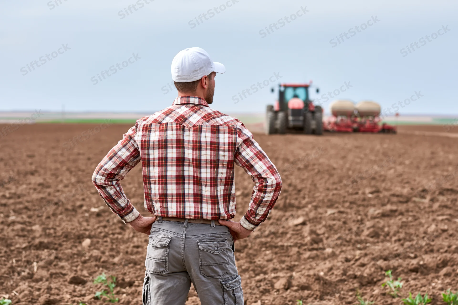 Farmer standing with hands on hips watching tractor plow field