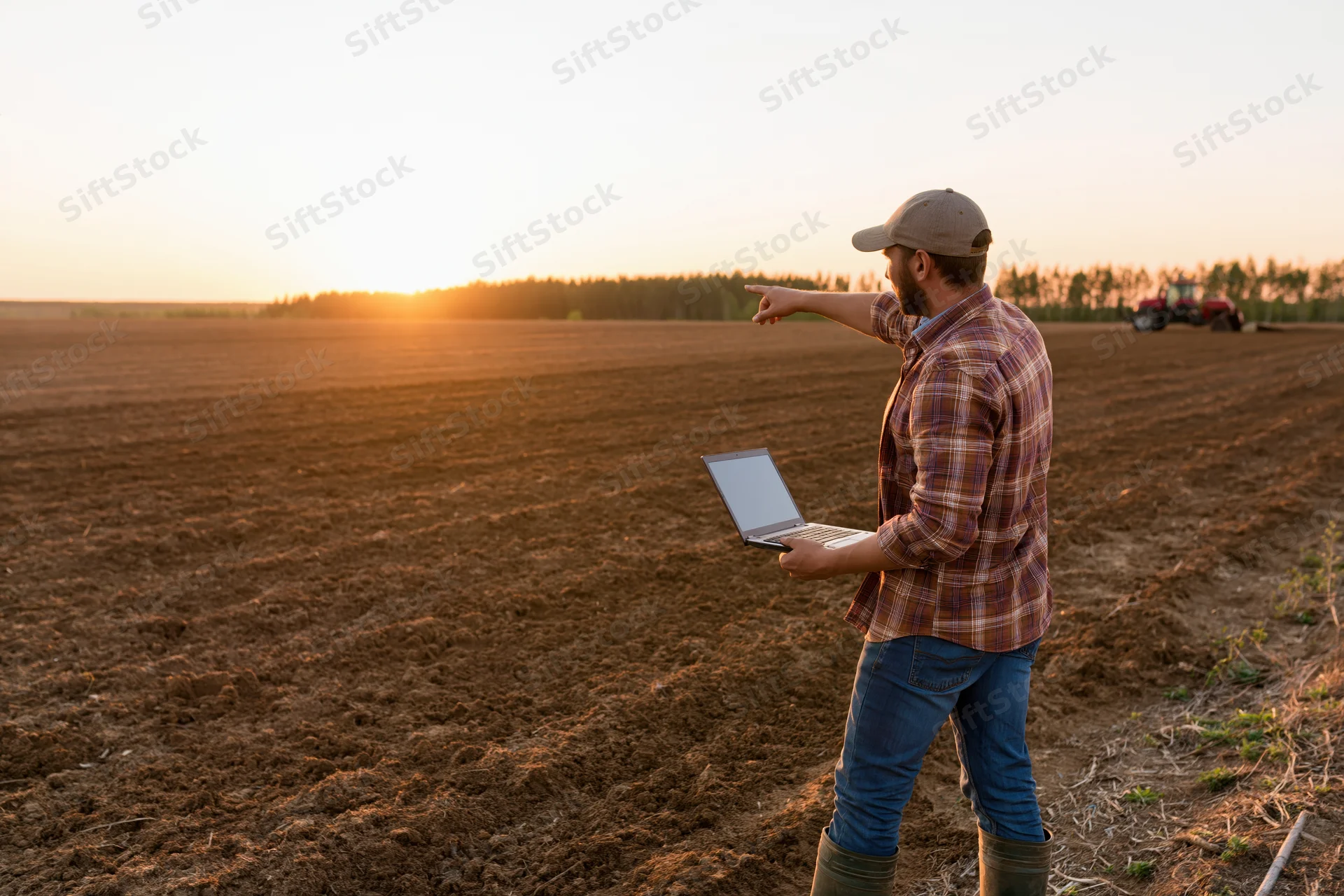 Farmer holding laptop and pointing across plowed field at sunset