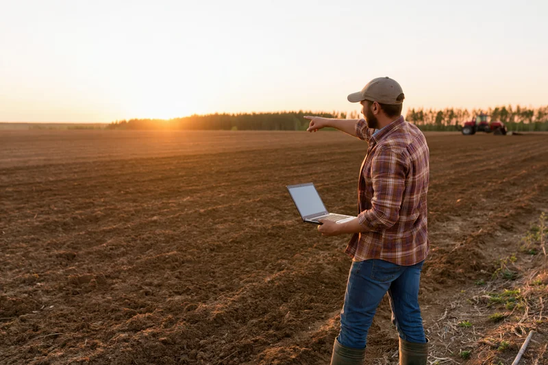 Farmer holding laptop and pointing across plowed field at sunset
