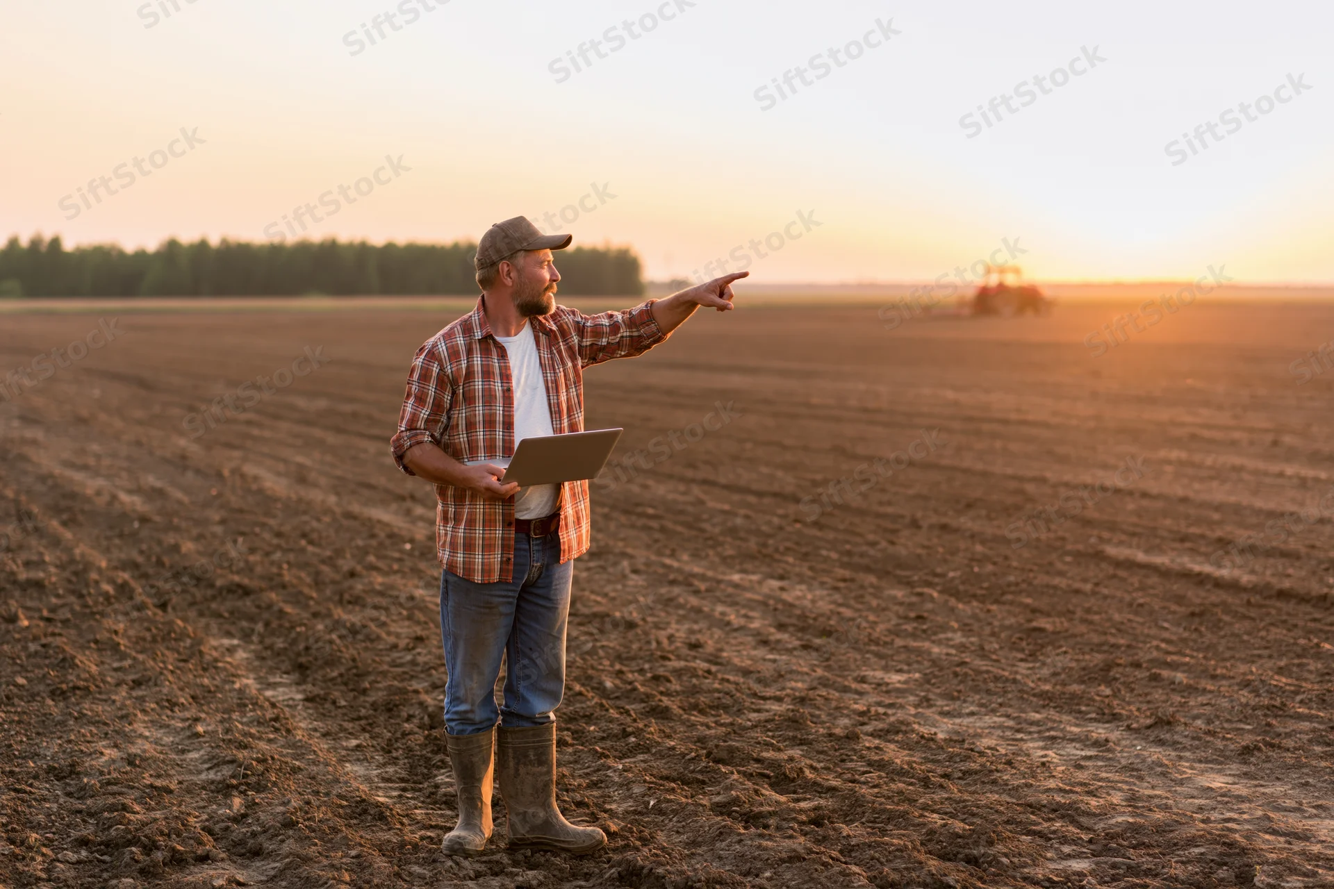 Farmer pointing across plowed field at tractor during sunset