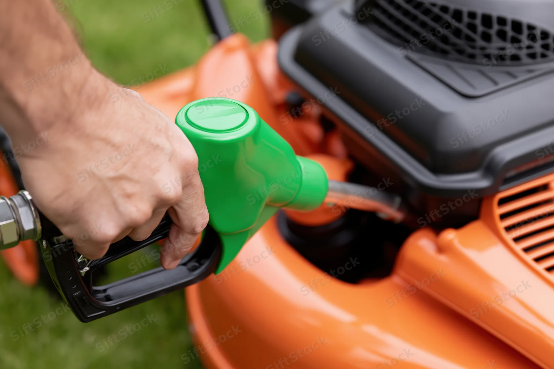 Hand refueling an orange lawnmower with a green nozzle on a grassy yard
