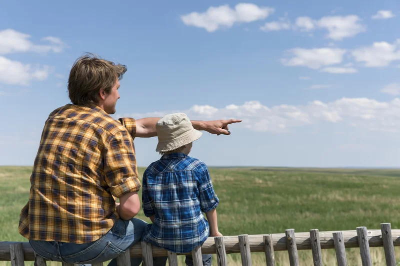 Father and son sitting on a wooden fence pointing at the countryside field