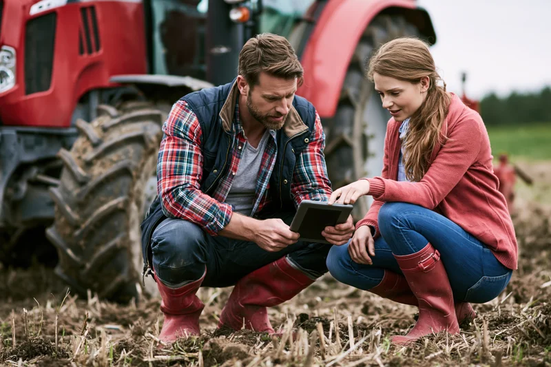 Man and woman crouching in a field examining soil with a tablet near a red tractor
