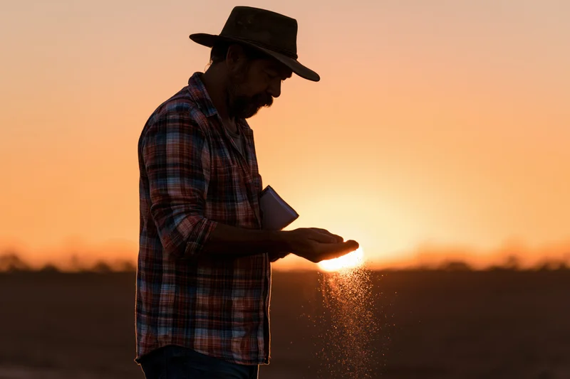 Farmer in plaid shirt and hat holding soil at sunset in rural field