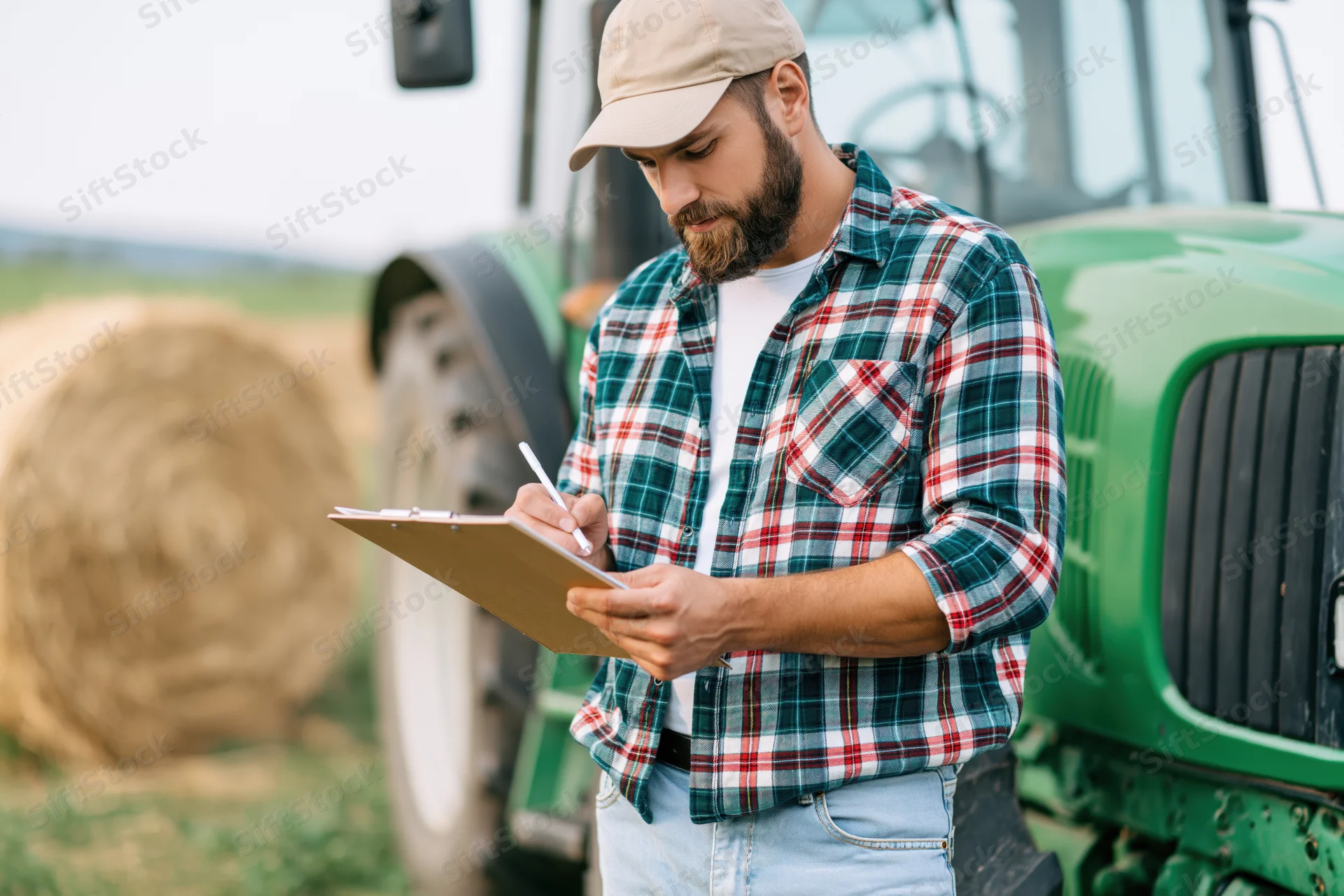 Farmer writing on clipboard in field beside green tractor and haybale