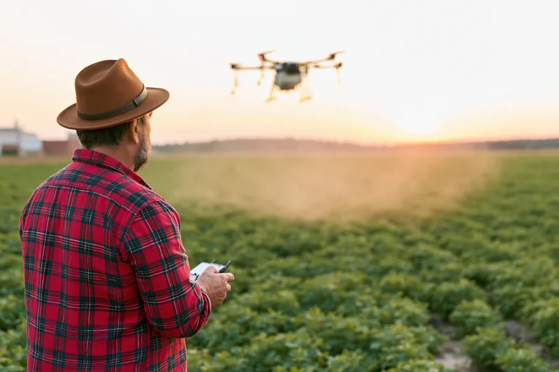 Man in red plaid shirt operating drone over crop field at sunset