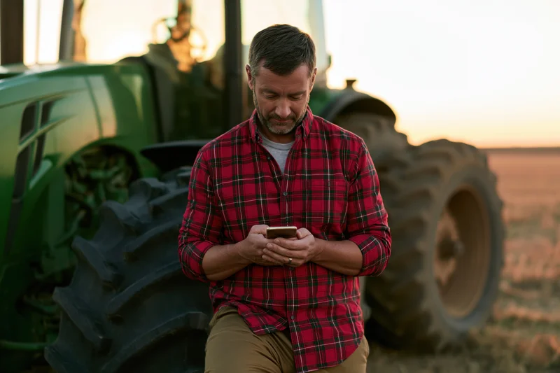 Man in red plaid shirt using smartphone beside green tractor in field at sunset