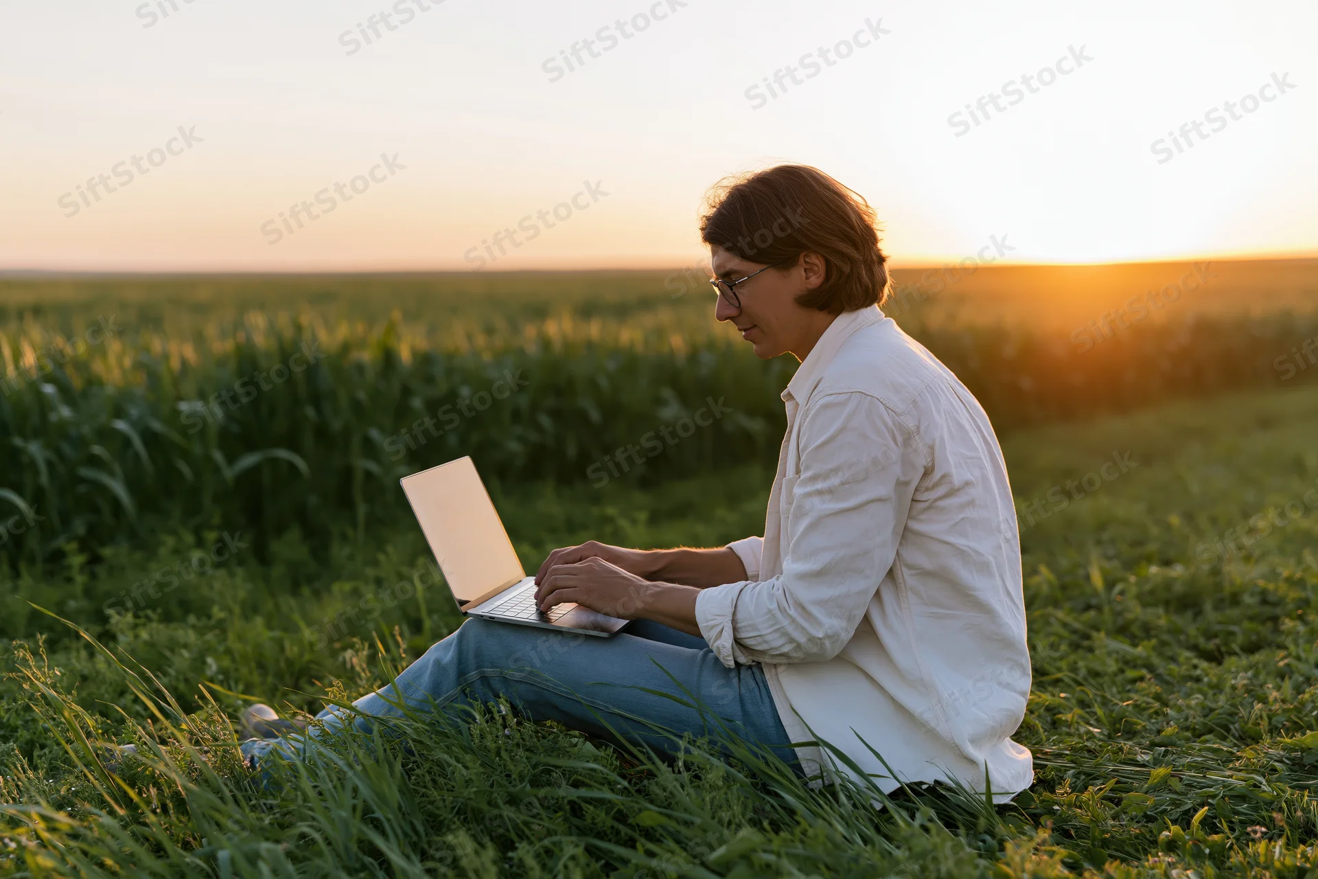 Woman typing on laptop while sitting in a grassy field at sunset