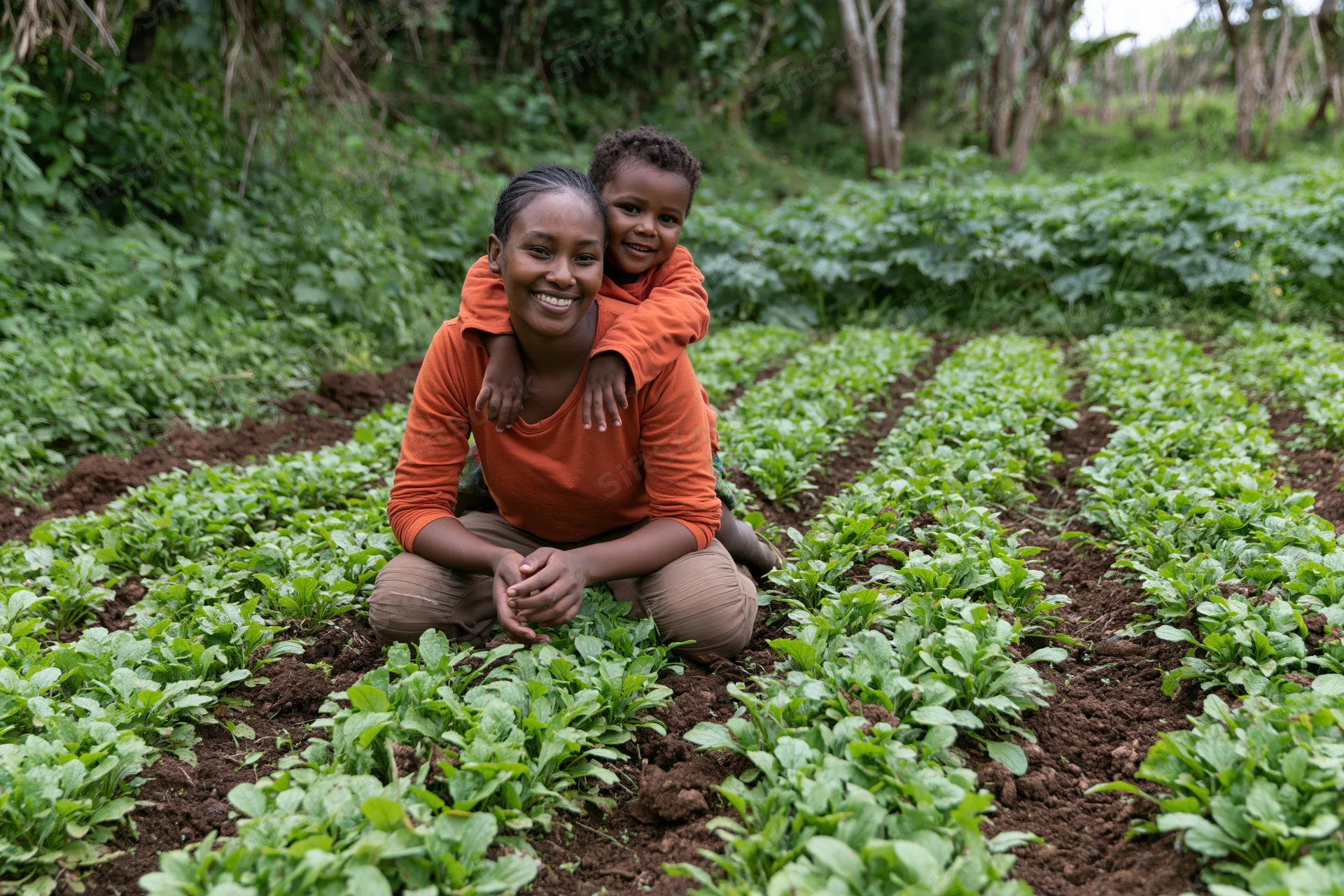 Woman crouching in vegetable garden with child on her back, surrounded by rows of leafy greens