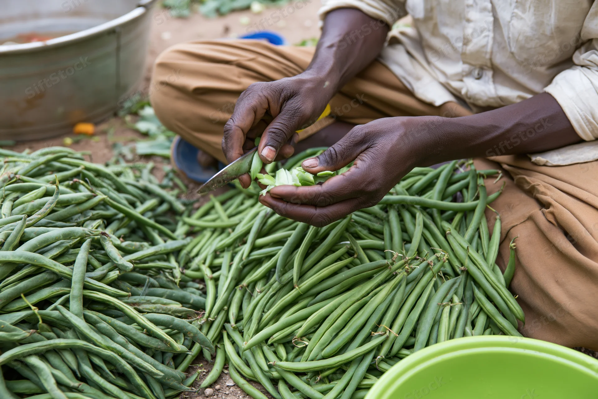 Man cutting green beans on ground outdoors with knife and pile of vegetables around him