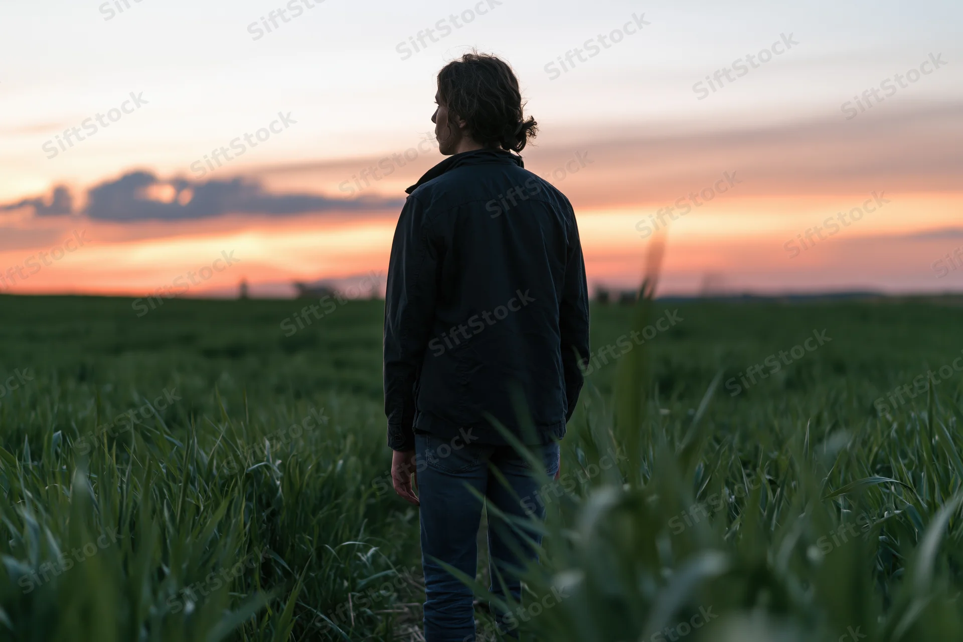 Man standing in a grassy field at sunset facing the horizon