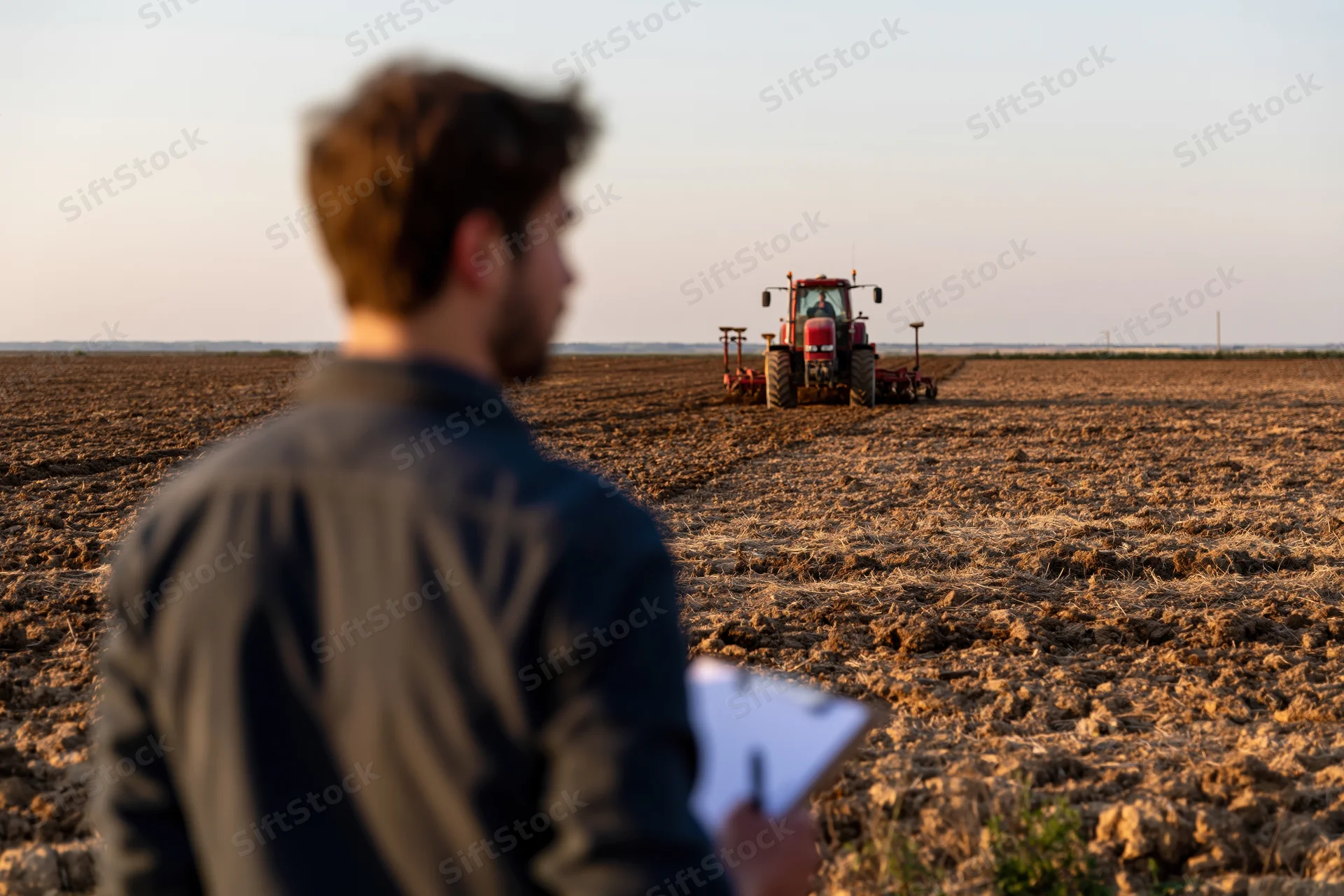 Farmer holding clipboard observing tractor plowing field at sunset