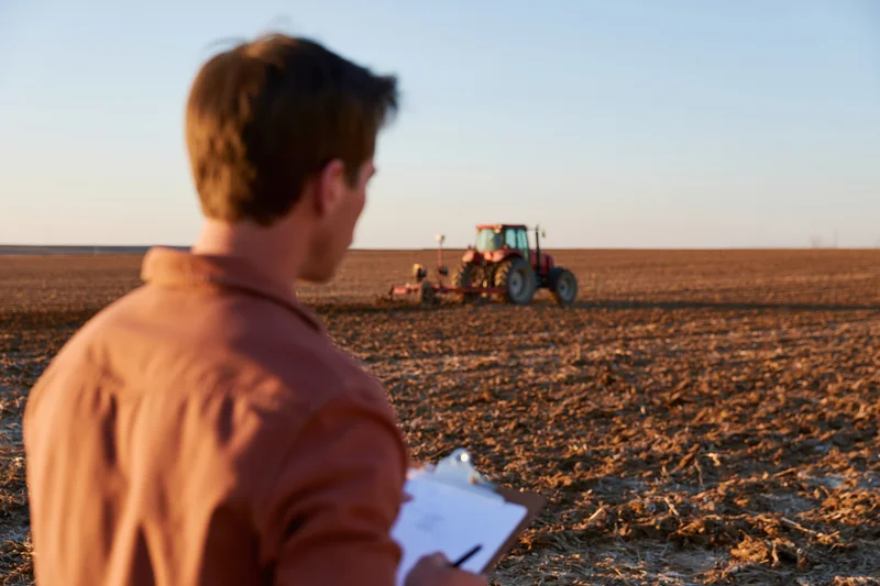 Young male farmer holding clipboard observing tractor plowing field at dusk
