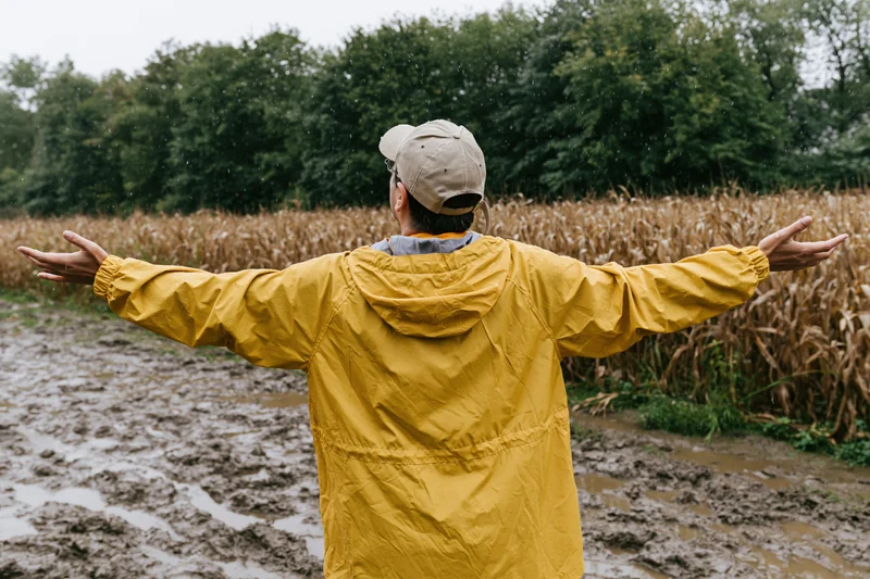 Man in yellow raincoat with arms outstretched standing in muddy cornfield during rain