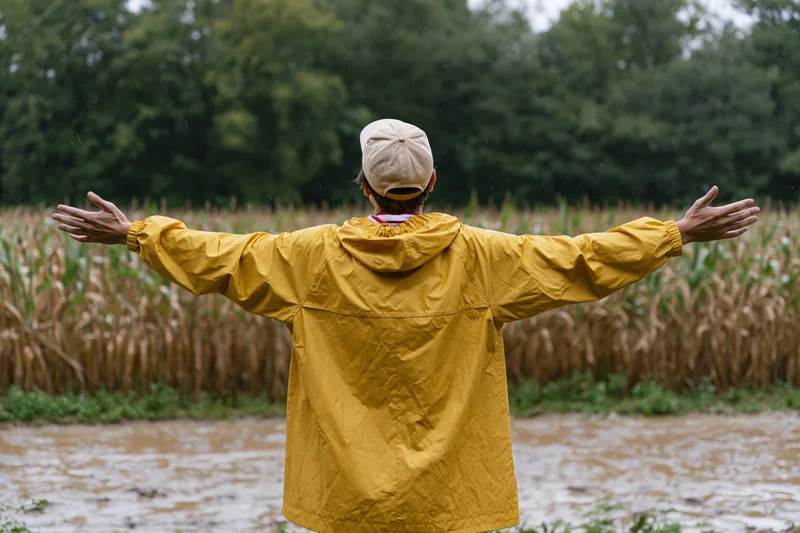 Man in yellow raincoat with arms outstretched standing in muddy field near cornfield during rain