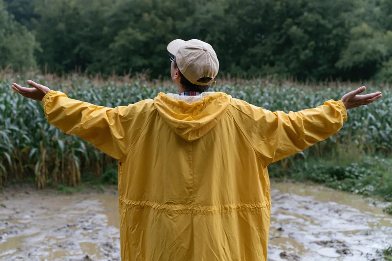 Person in yellow raincoat with arms outstretched standing in muddy field near corn crop