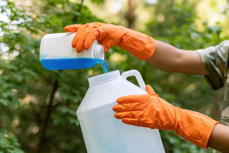 Woman in orange gloves pouring blue liquid from a bottle into a white jug outdoors in a garden