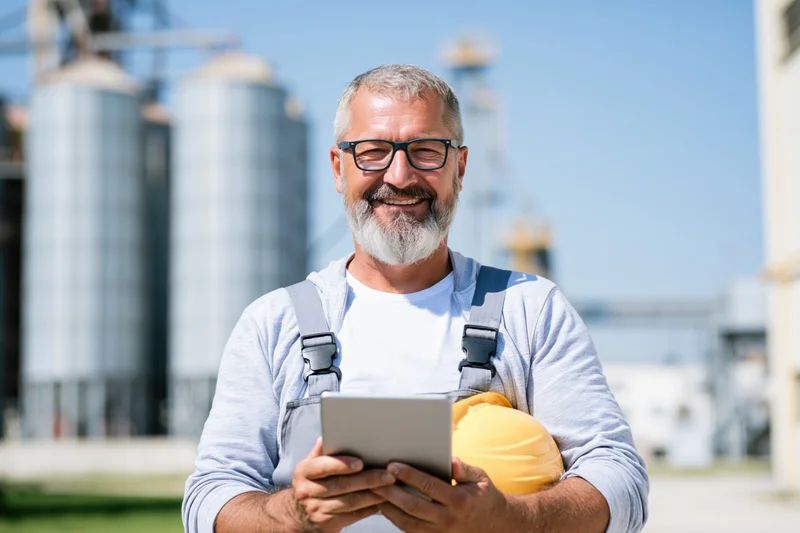Smiling man in overalls holding tablet and hardhat at industrial facility