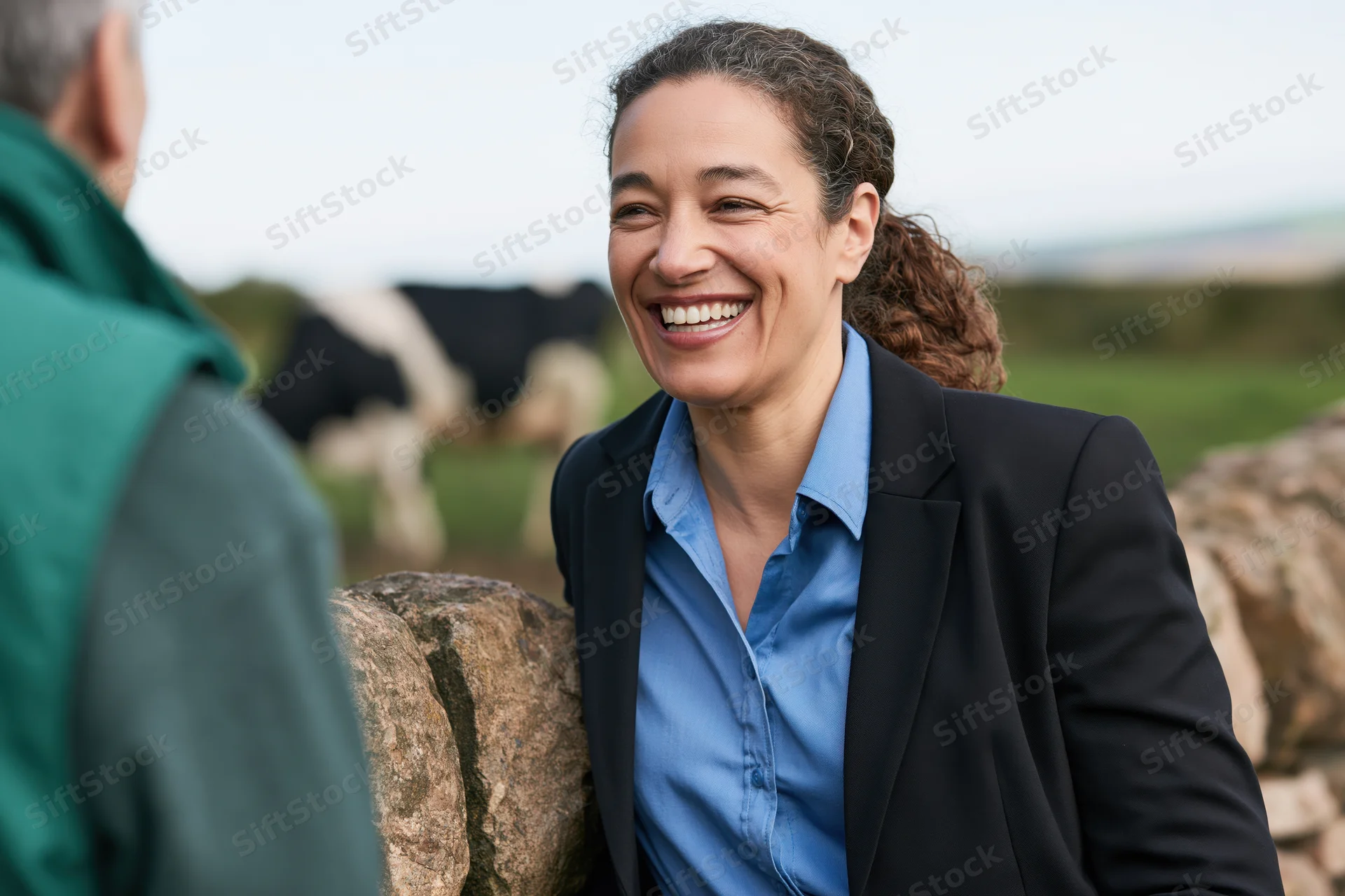 Woman in suit smiling while talking to a man by a stone wall in a pasture with cows grazing in the background