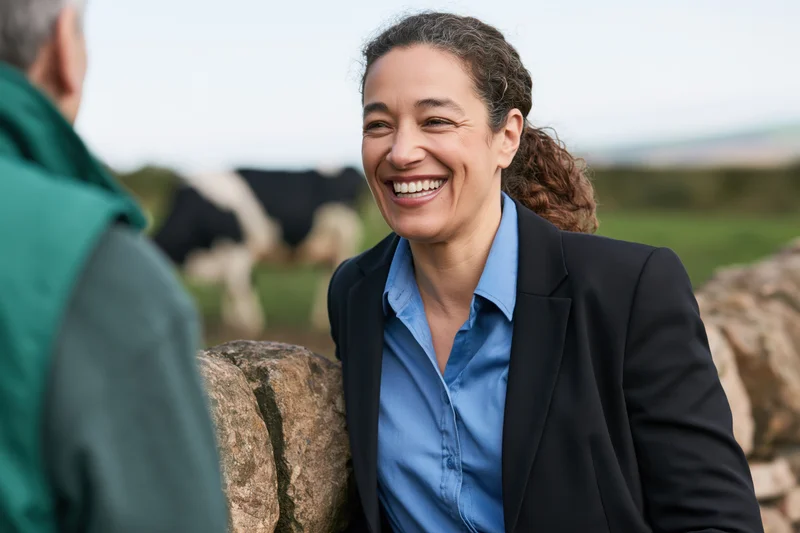 Woman in suit smiling while talking to a man by a stone wall in a pasture with cows grazing in the background