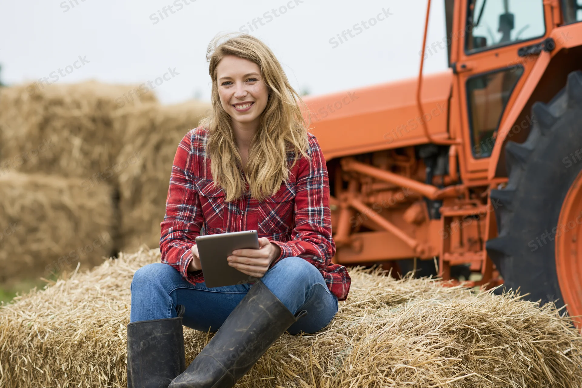 Woman sitting on hay bale holding tablet with tractor in background