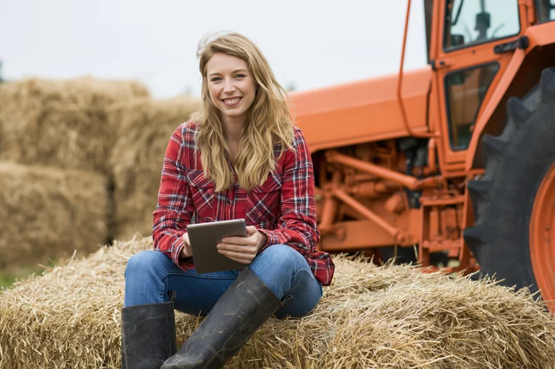 Woman sitting on hay bale holding tablet with tractor in background