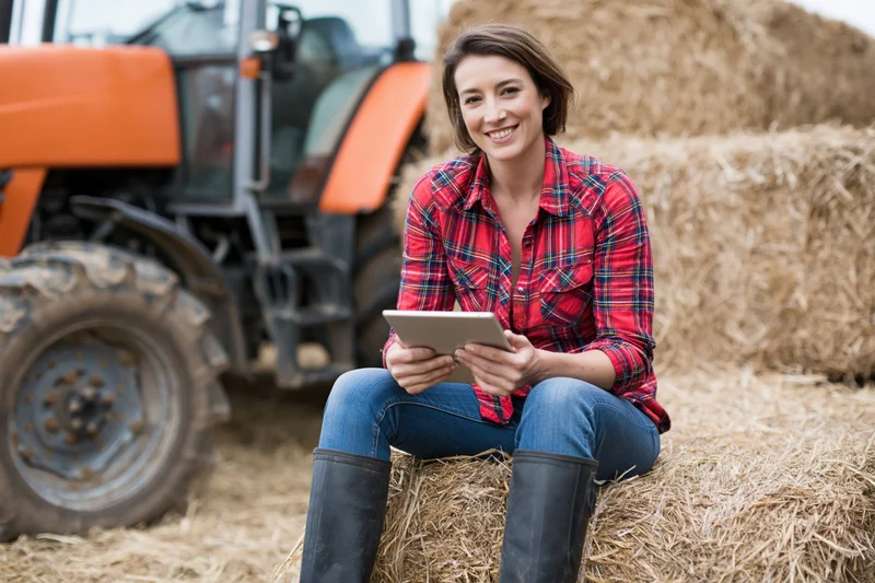 Woman farmer sitting on haybale using tablet in field near tractor