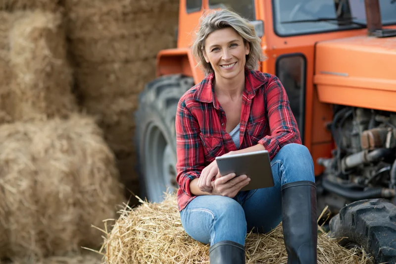 Woman sitting on hay bale holding tablet near orange tractor in rural field