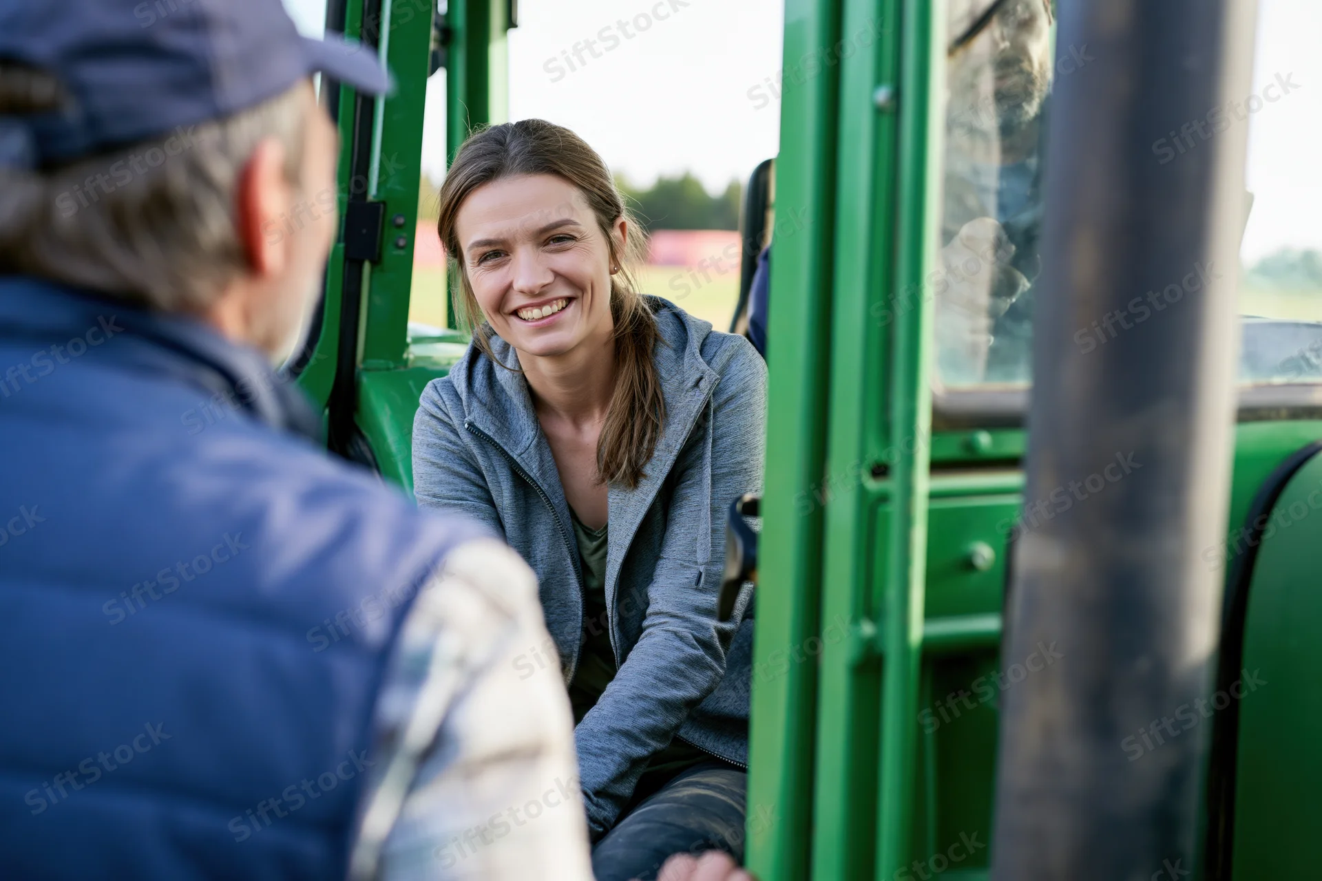 Woman smiling while sitting in tractor cab talking to male farmer outdoors