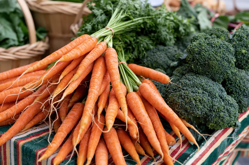 Bunches of fresh carrots and broccoli displayed on a striped cloth at a farmers market stall