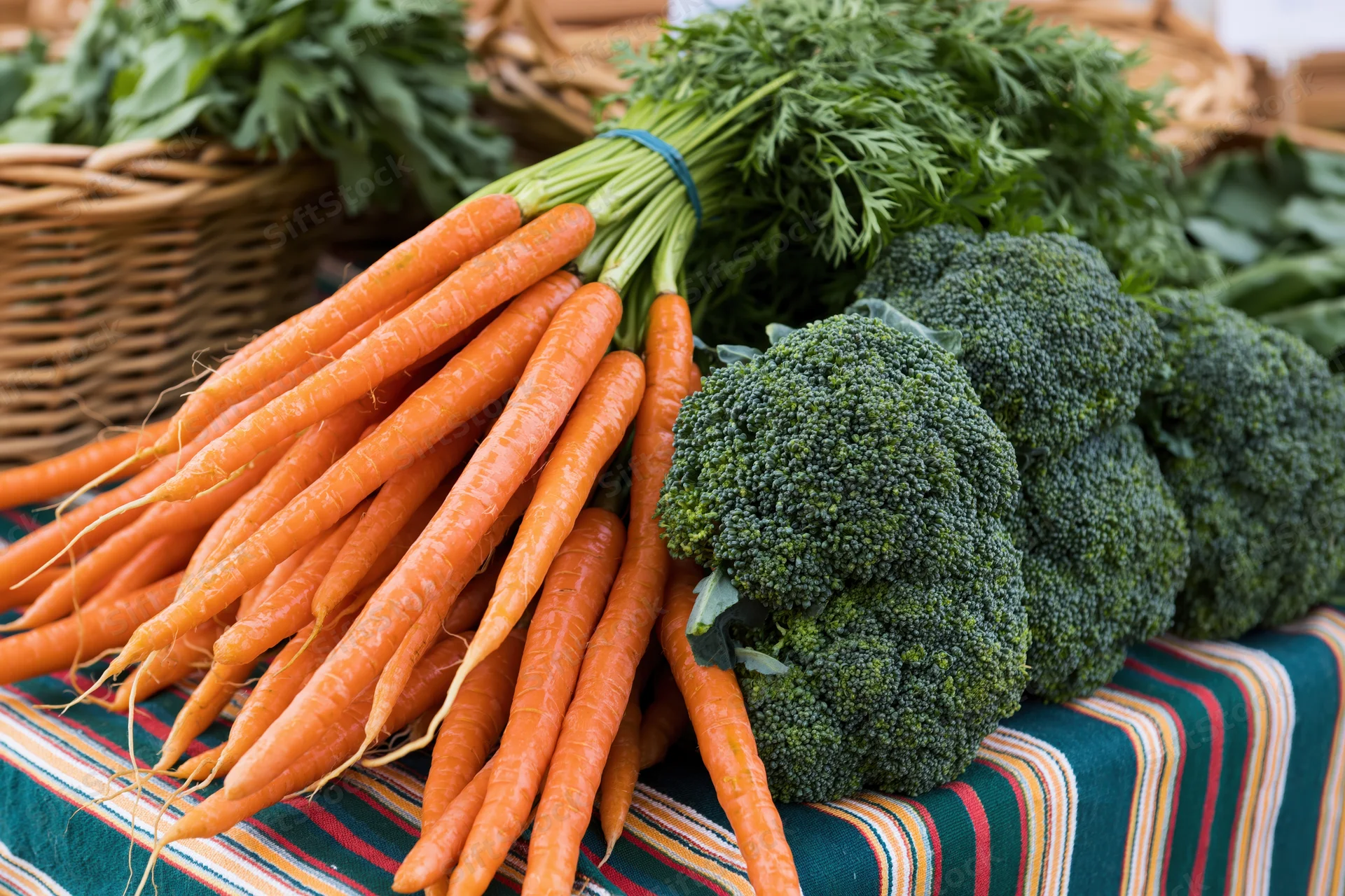 Bunches of fresh carrots and broccoli displayed on a striped cloth at a farmers market stall