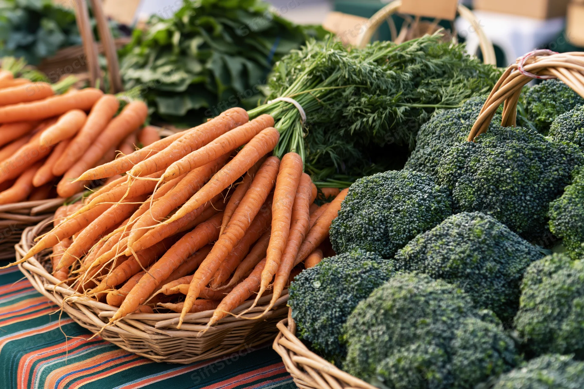 Baskets of fresh carrots and broccoli arranged on a striped cloth at a farmers market stall