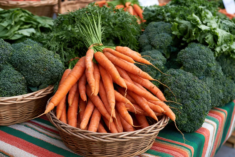 Carrots and broccoli in wicker baskets on a striped tablecloth at a farmers market