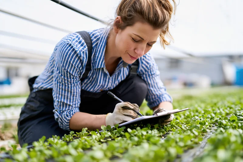 Woman crouching in greenhouse writing notes on clipboard among leafy plants